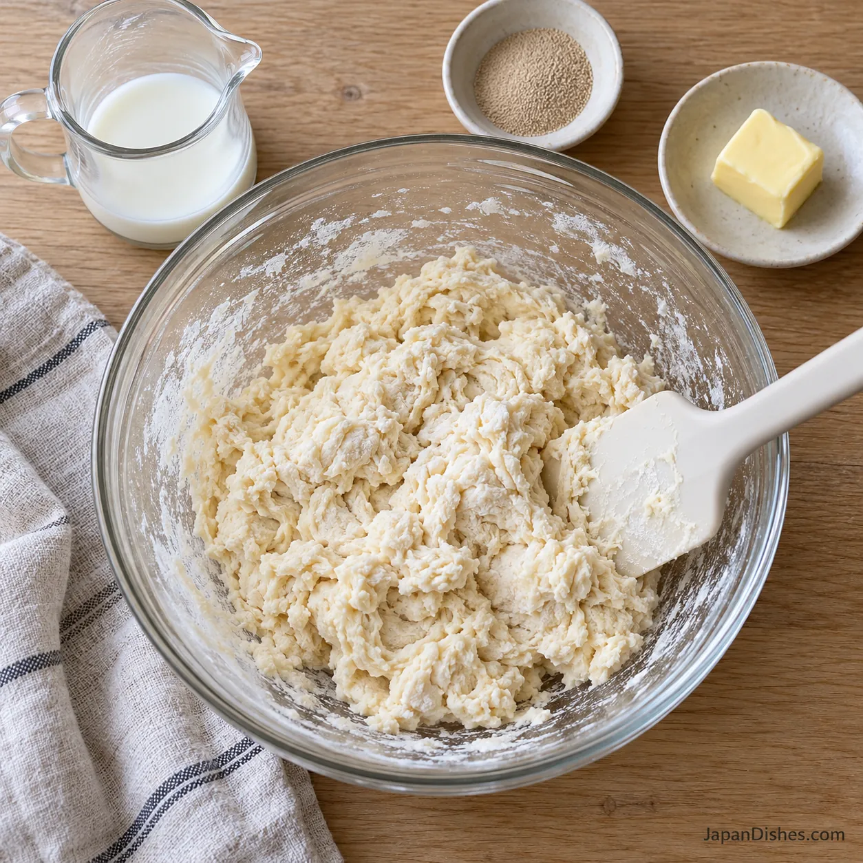 Shaggy milk bread dough in a glass bowl with a spatula and butter set aside